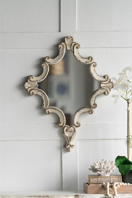 The Artistic Diamond Scrollwork Mirror in Antique White hangs on a white paneled wall above stacked books, coral, a green leaf, and a white orchid in a glass vase. Perfect as a decorative accent for your living room, entryway, bedroom, or office.