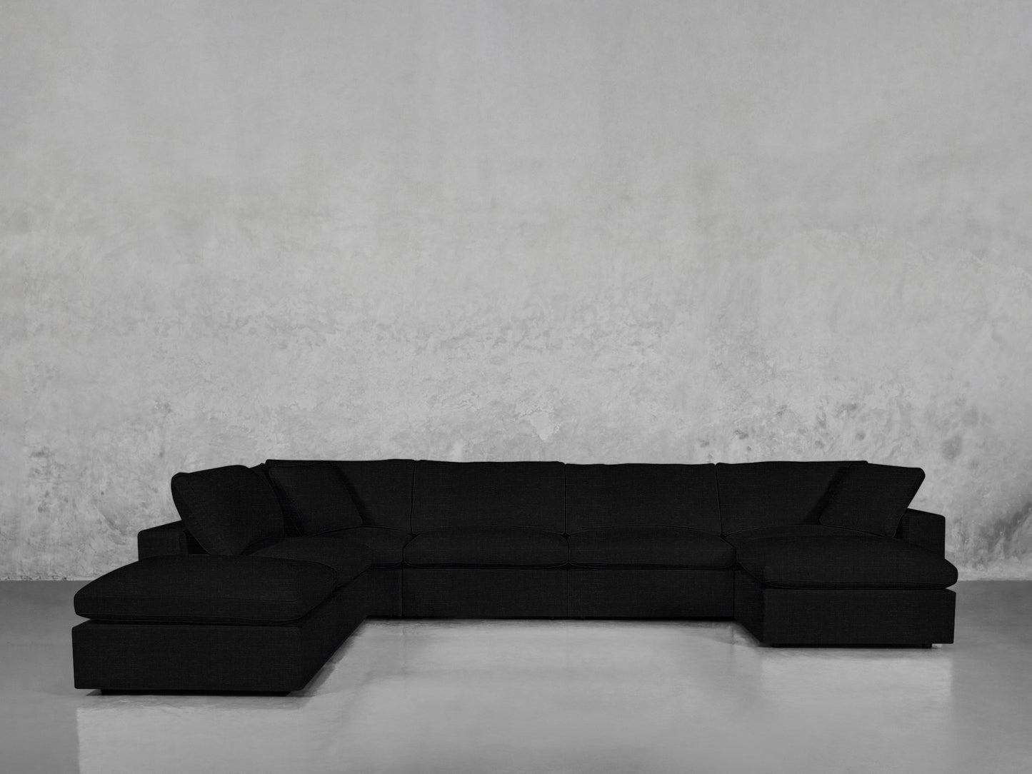 The 7-Seat Modular Chaise Corner Lounger Sectional, a large black modern sofa with deep cushions, is set against a plain textured gray wall on a smooth reflective floor.