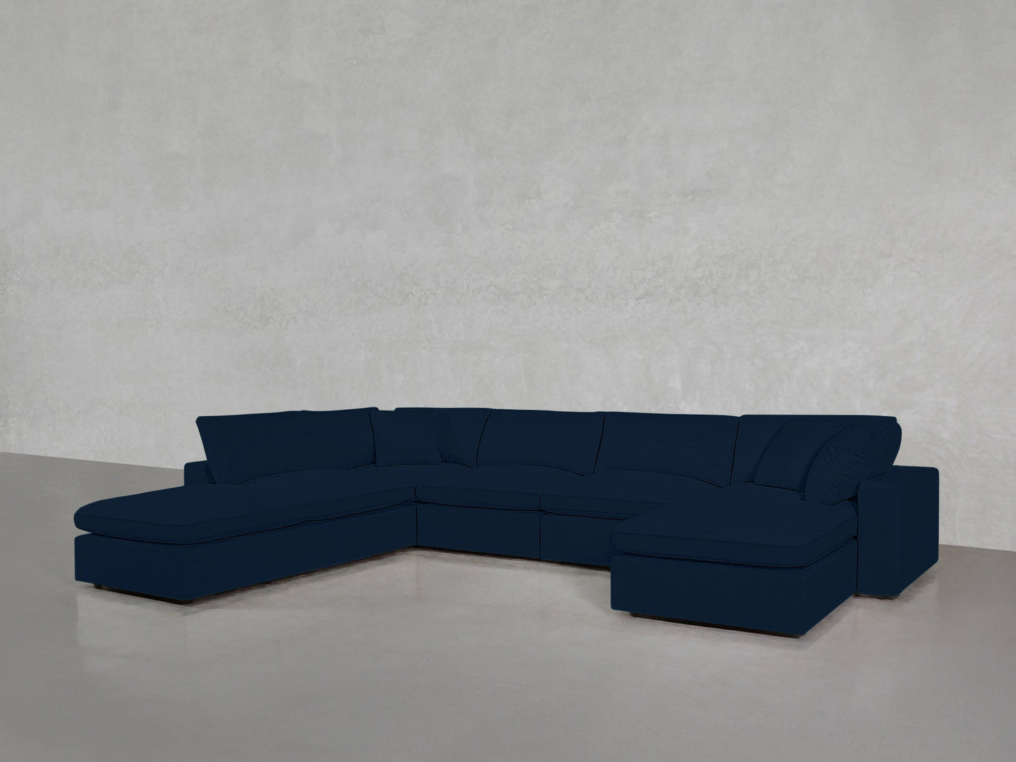 A 7-Seat Modular Chaise Corner Lounger Sectional with clean lines, upholstered in dark blue, is arranged in an L-shape against a light gray textured wall on a polished concrete floor.