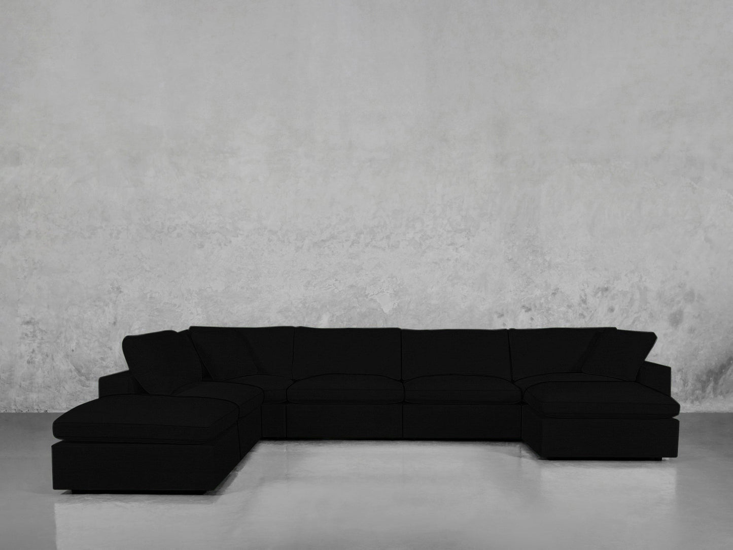 A spacious black 7-Seat Modular Chaise Corner Lounger Sectional with a left-side chaise rests against a plain, light gray concrete wall and a smooth, reflective floor.