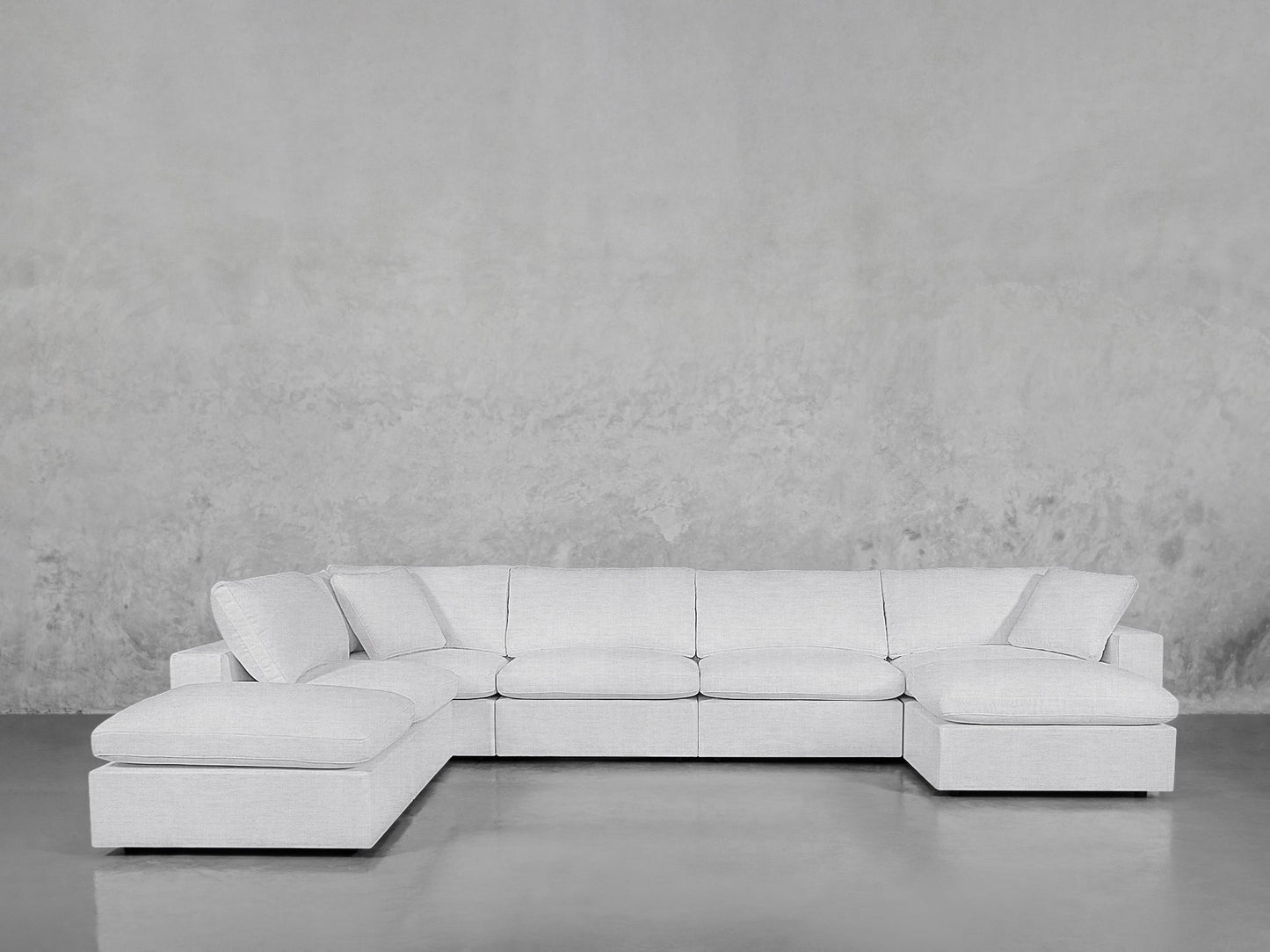 The 7-Seat Modular Chaise Corner Lounger Sectional in light gray, with plush seat and back cushions, wide armrests, and two corner pieces, sits against a textured gray wall on a polished concrete floor.