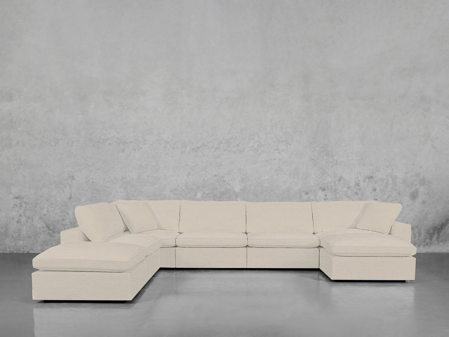 The 7-Seat Modular Chaise Corner Lounger Sectional, in light beige with plush cushions, is set against a textured gray wall on a glossy concrete floor.