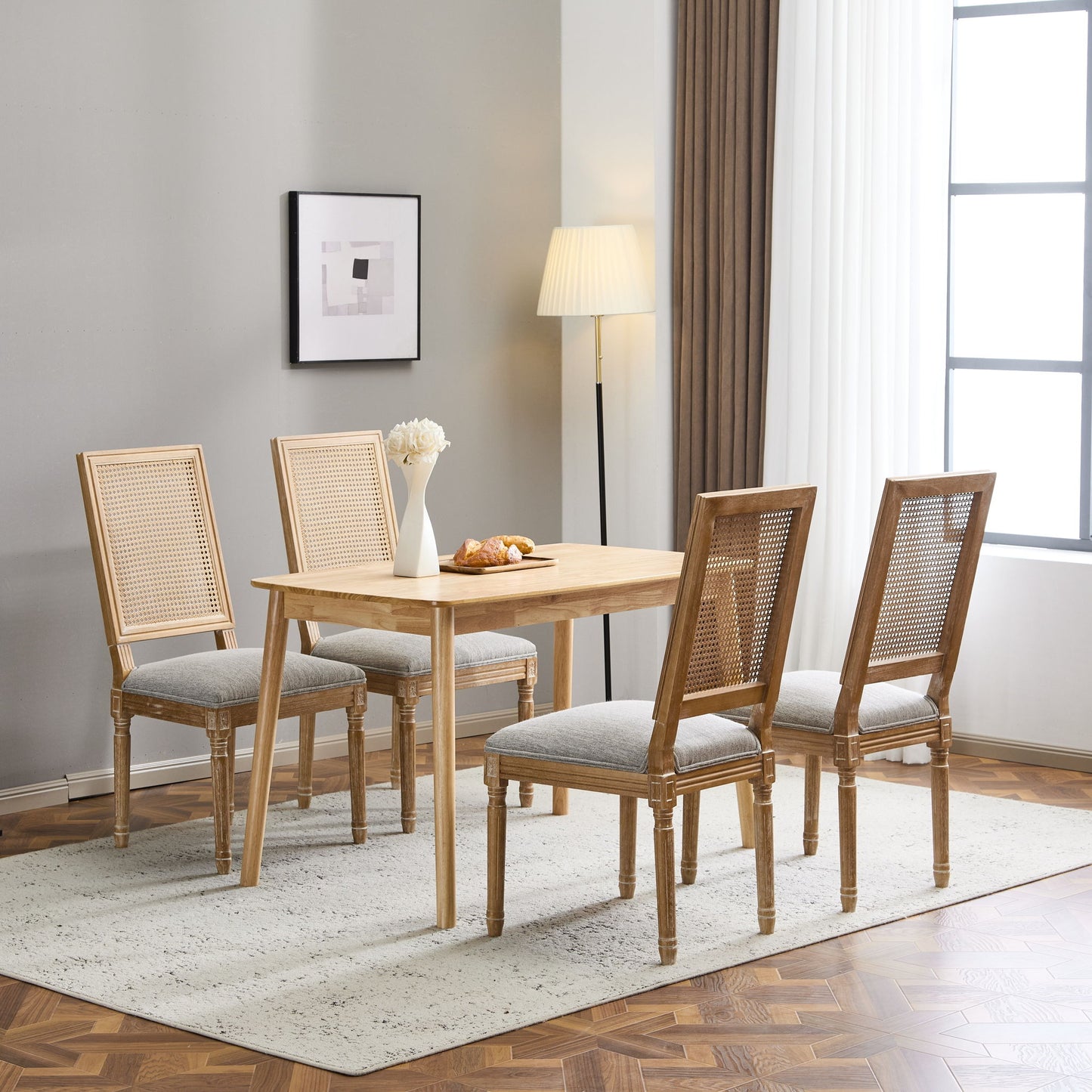 A dining room with a wooden table, four French Country Dining Chairs with rectangular cane backrests and smooth upholstered seats, a white vase of flowers, pastries on a plate, a floor lamp, abstract art, and a large window with white curtains.