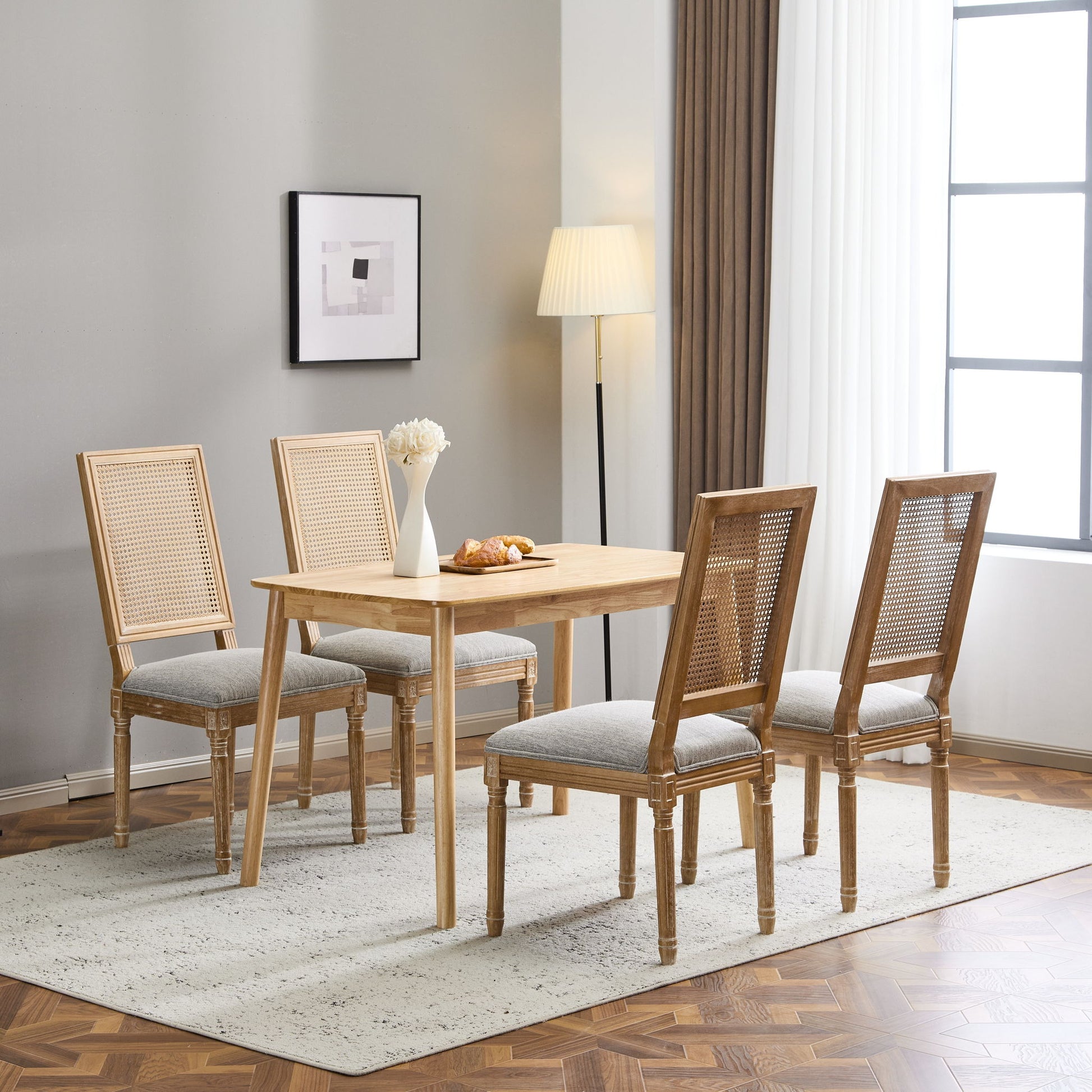 A dining room with a wooden table, four French Country Dining Chairs with rectangular cane backrests and smooth upholstered seats, a white vase of flowers, pastries on a plate, a floor lamp, abstract art, and a large window with white curtains.