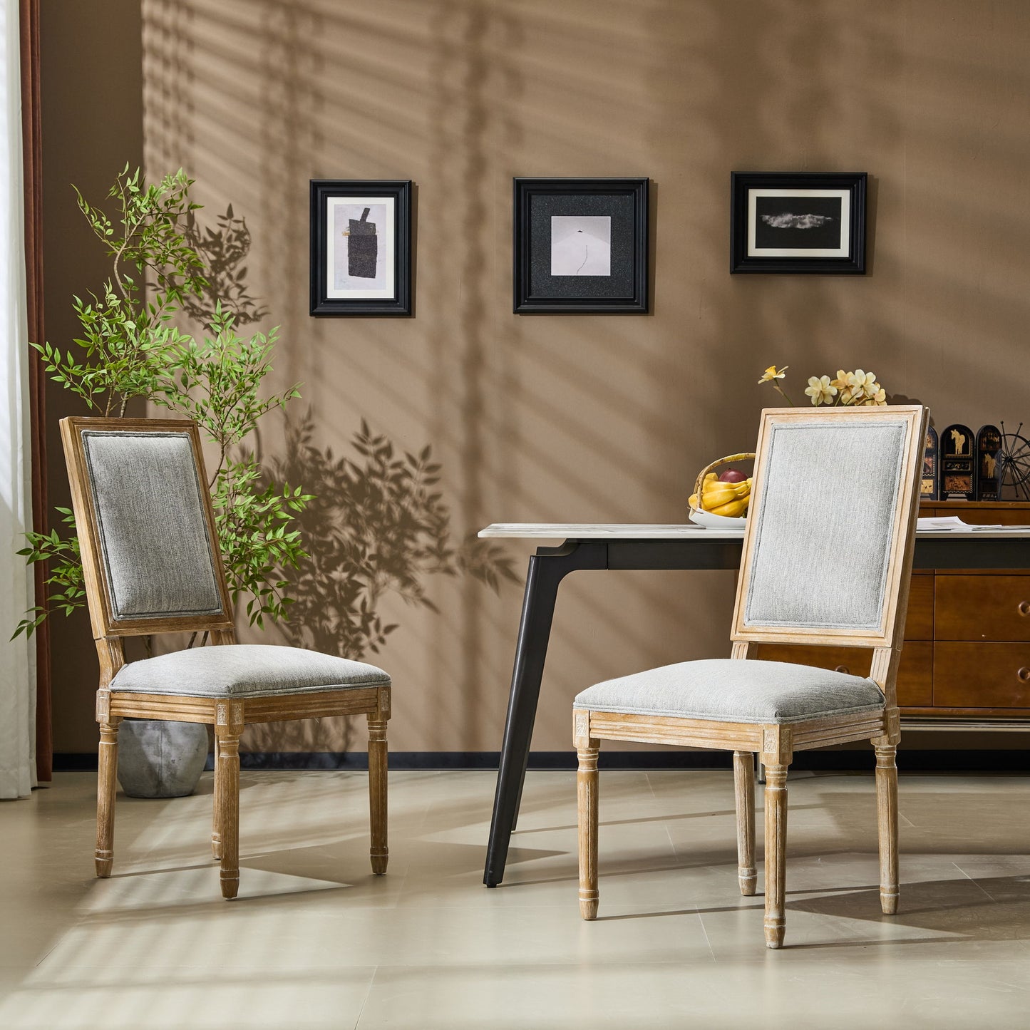 Two French Country Style Dining Chairs with upholstered seats and fluted legs are set in a modern room with a brown wall, three small framed pictures above, a desk with fruit and decor against the wall, and a green plant nearby.