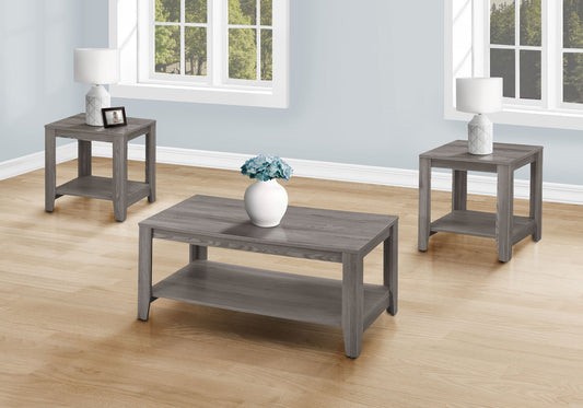 A living room with light wood floors features a gray transitional table set (coffee table and two side tables). Each side table has a white lamp, and the coffee table holds a white vase with blue flowers. Sunlight streams through the windows.