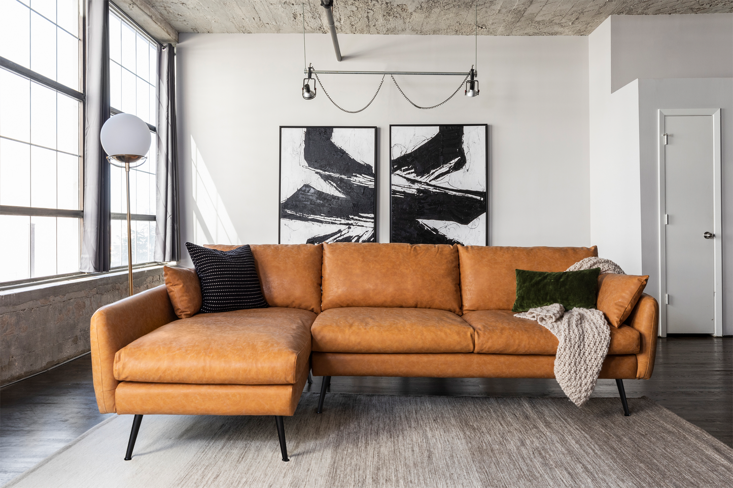 A modern living room featuring a Park Sectional Sofa in brown leather, adorned with black and white abstract art on the wall, illuminated by a floor lamp, and bathed in natural light from large windows. A knitted blanket and pillows enhance the comfort of the setting. The wooden floor is partially covered by a light rug.