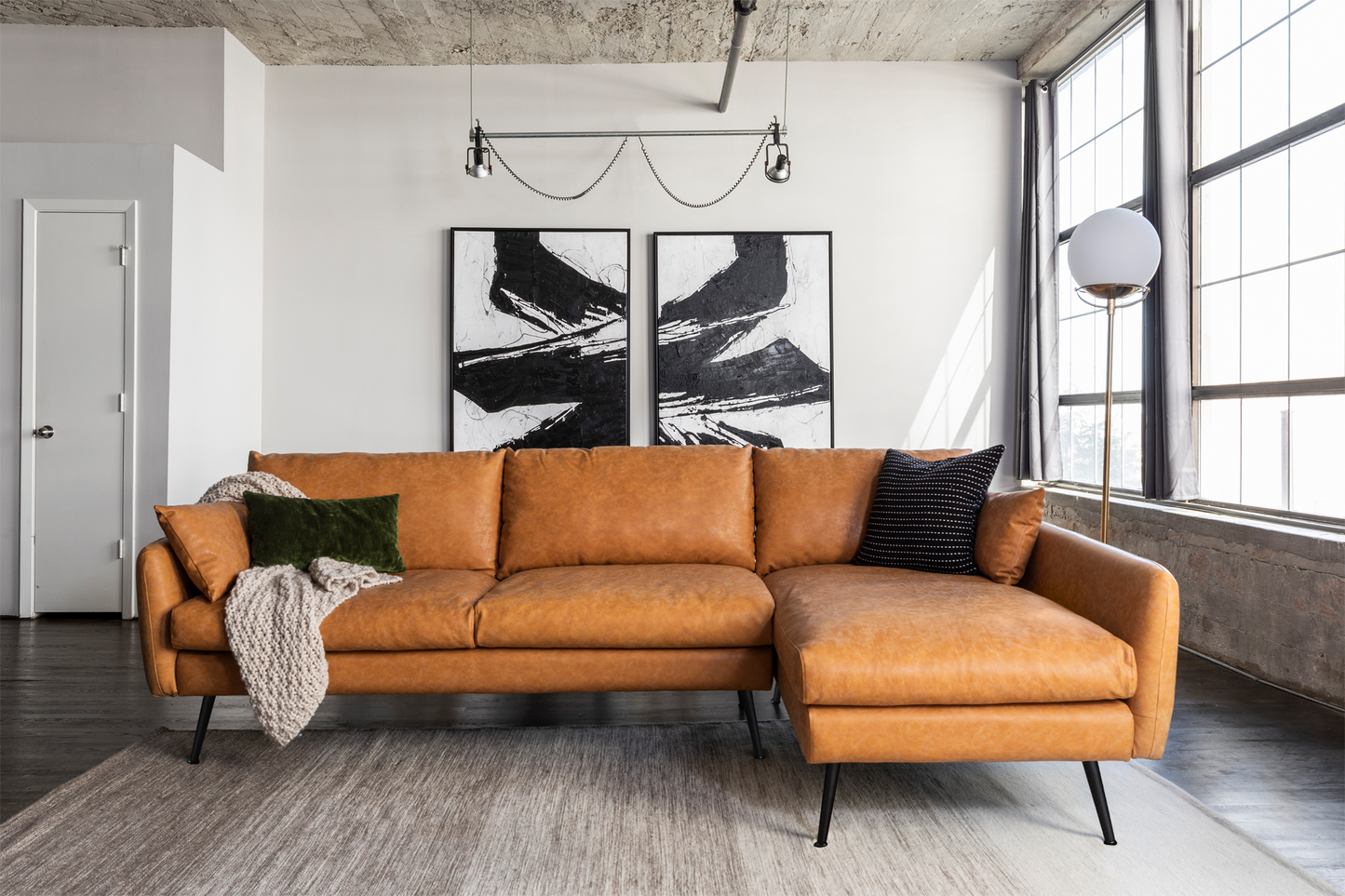 A contemporary living room showcases a spacious Park Sectional Sofa in brown leather, enhanced with green and black pillows and a beige throw. Behind the sofa, two abstract black and white paintings are displayed. The room is brightly illuminated by large windows.
