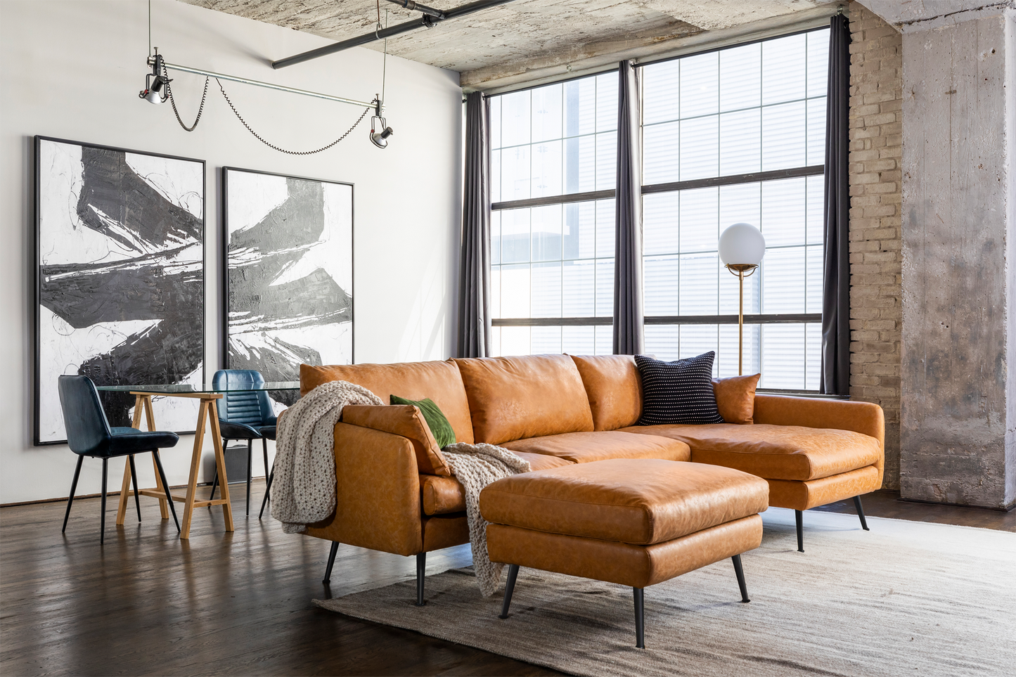 In a modern living room, there's a spacious Park Sectional Sofa paired with a matching ottoman on top of an elegantly placed rug. Two abstract black and white paintings adorn the wall. Nearby, there's a small dining table accompanied by chairs, while large windows with curtains fill the space with natural light.