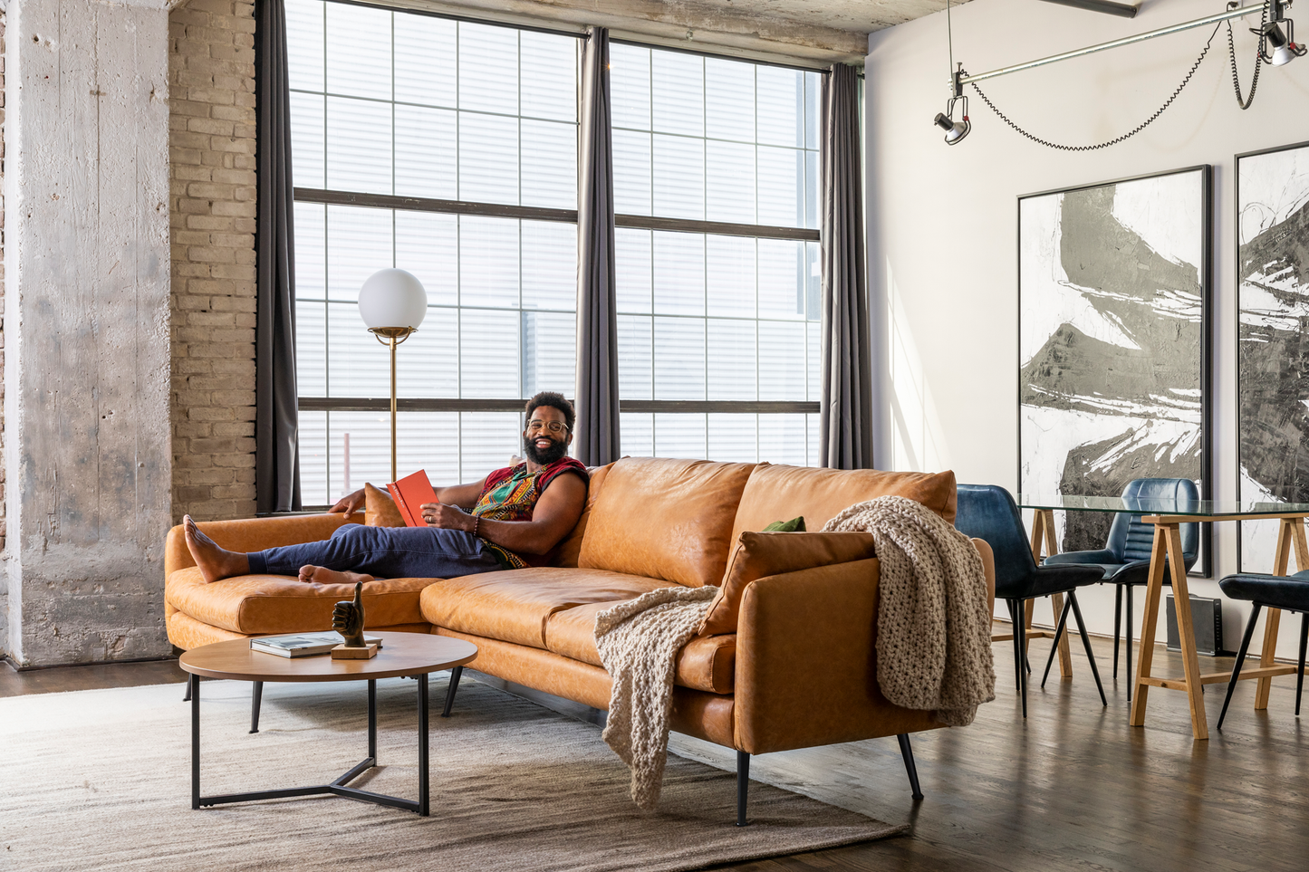 A person relaxes on a spacious Park Sectional Sofa made of tan leather in a bright loft apartment with tall windows. The room is decorated with abstract art, features a round coffee table, a knitted blanket, and includes a dining area with dark blue chairs.