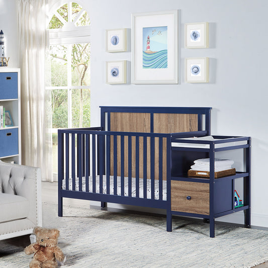 The Connelly 4-in-1 Crib and Changer Combo in navy blue and wood stands in a bright nursery with framed lighthouse art, a window with white curtains, and a teddy bear on a light gray rug.