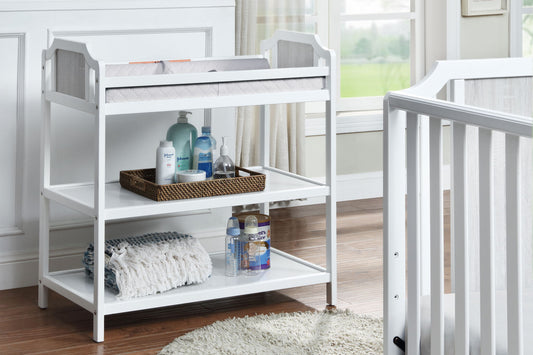 The Brees - Changing Table, finished in white, features shelves stocked with bottles, lotion, a basket, and folded blankets. It is placed next to a white crib in a bright, softly lit nursery.