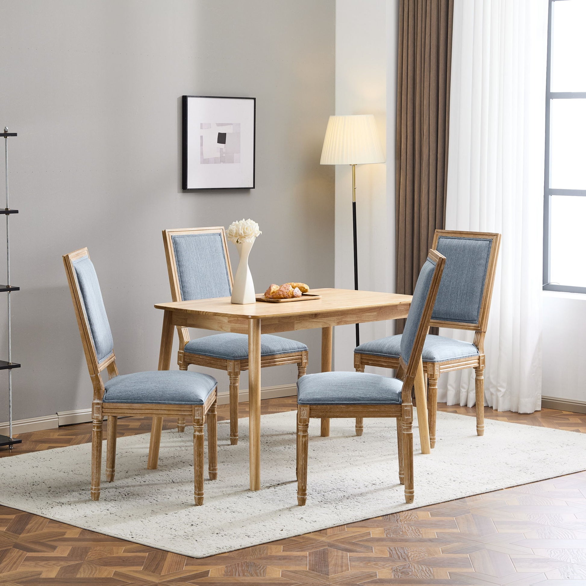 A wooden dining table with four French Country Style Dining Chairs featuring upholstered seating and fluted legs sits on a light rug. On the table are a vase of white flowers and bread, with a floor lamp, framed art, and large window in the background.