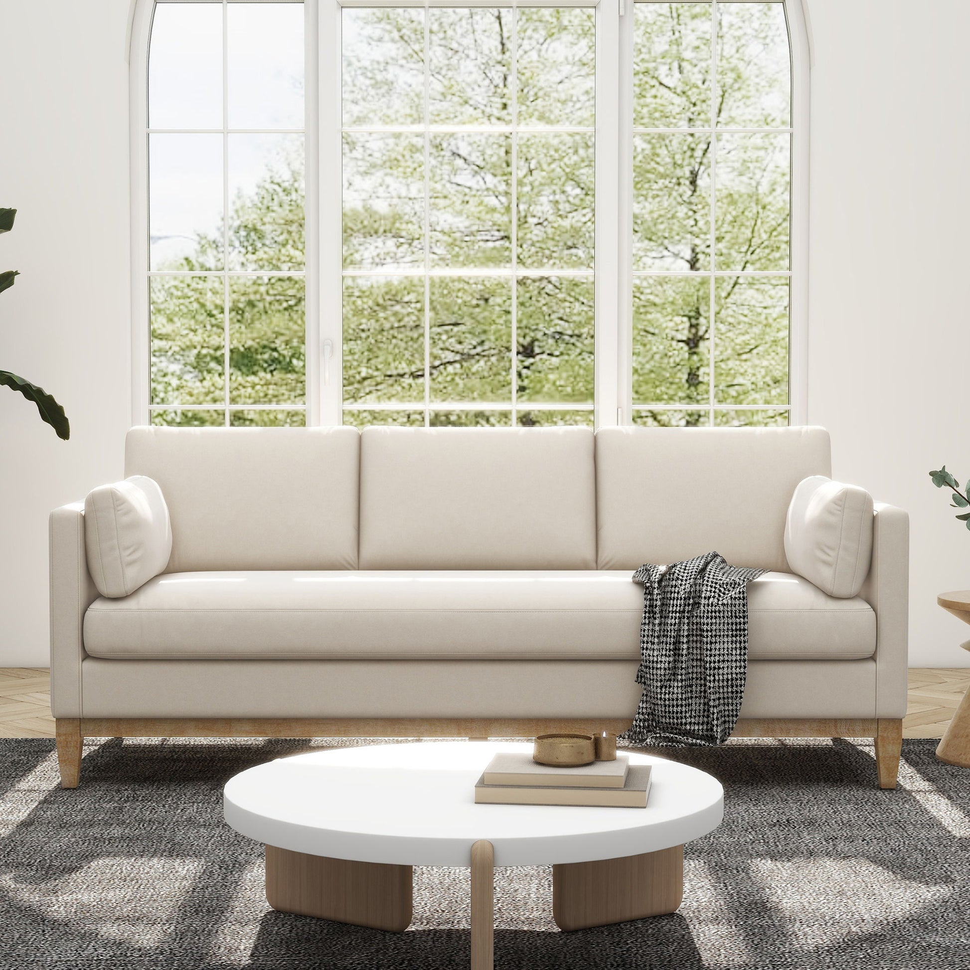 A modern living room with the Knox - Modern Farmhouse Sofa in light beige, a black and white throw, a round white coffee table, and a large window revealing green trees. The cozy space features minimalist design elements.