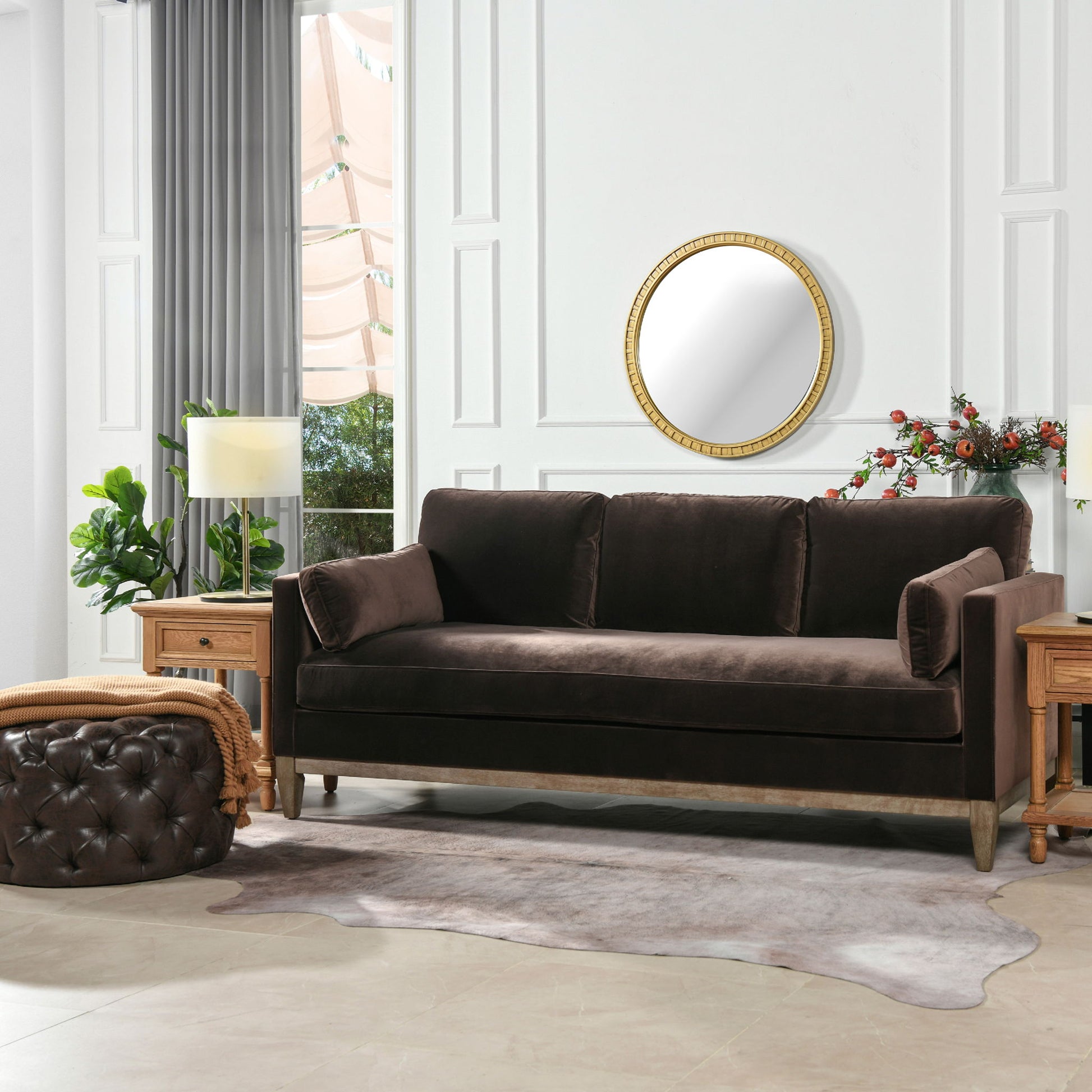 A modern living room featuring the Knox - Modern Farmhouse Sofa in dark brown velvet, two wooden side tables with lamps, a round mirror on a white paneled wall, green plants, a tufted ottoman, and a cowhide rug.
