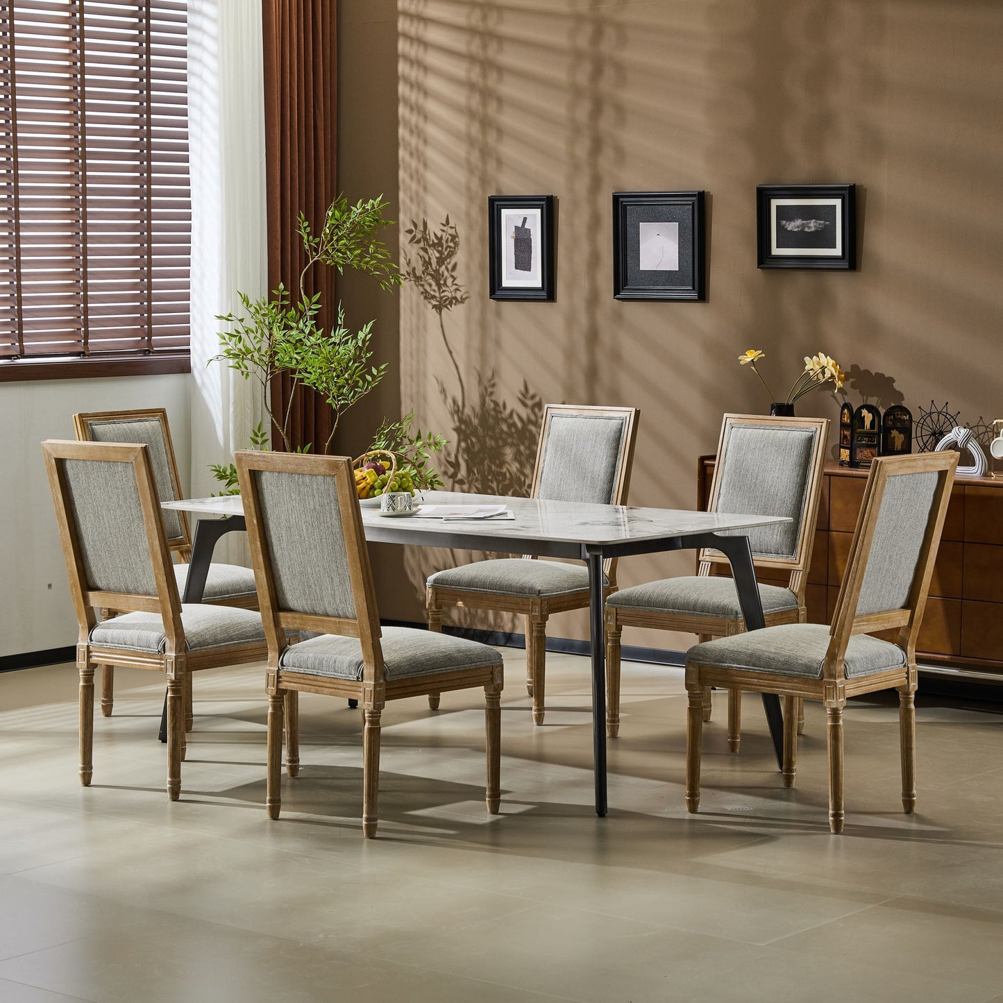 A dining room features a rectangular table surrounded by six French Country Style Dining Chairs with upholstered seating and fluted legs, set against beige walls with framed art, a potted plant, and sunlight streaming through window blinds.