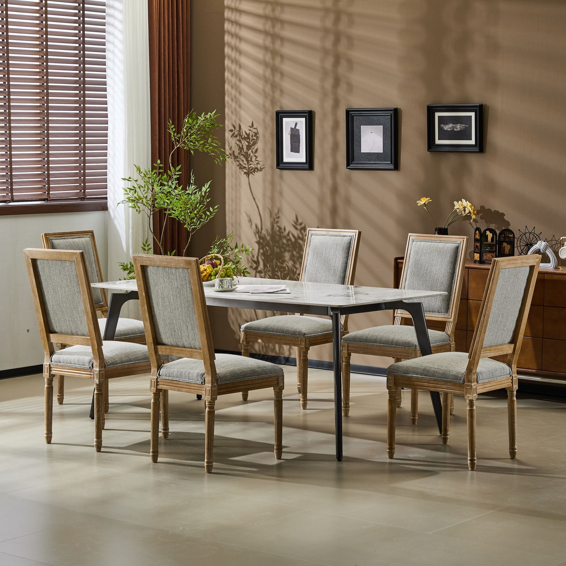 A dining room features a rectangular table surrounded by six French Country Style Dining Chairs with upholstered seating and fluted legs, set against beige walls with framed art, a potted plant, and sunlight streaming through window blinds.