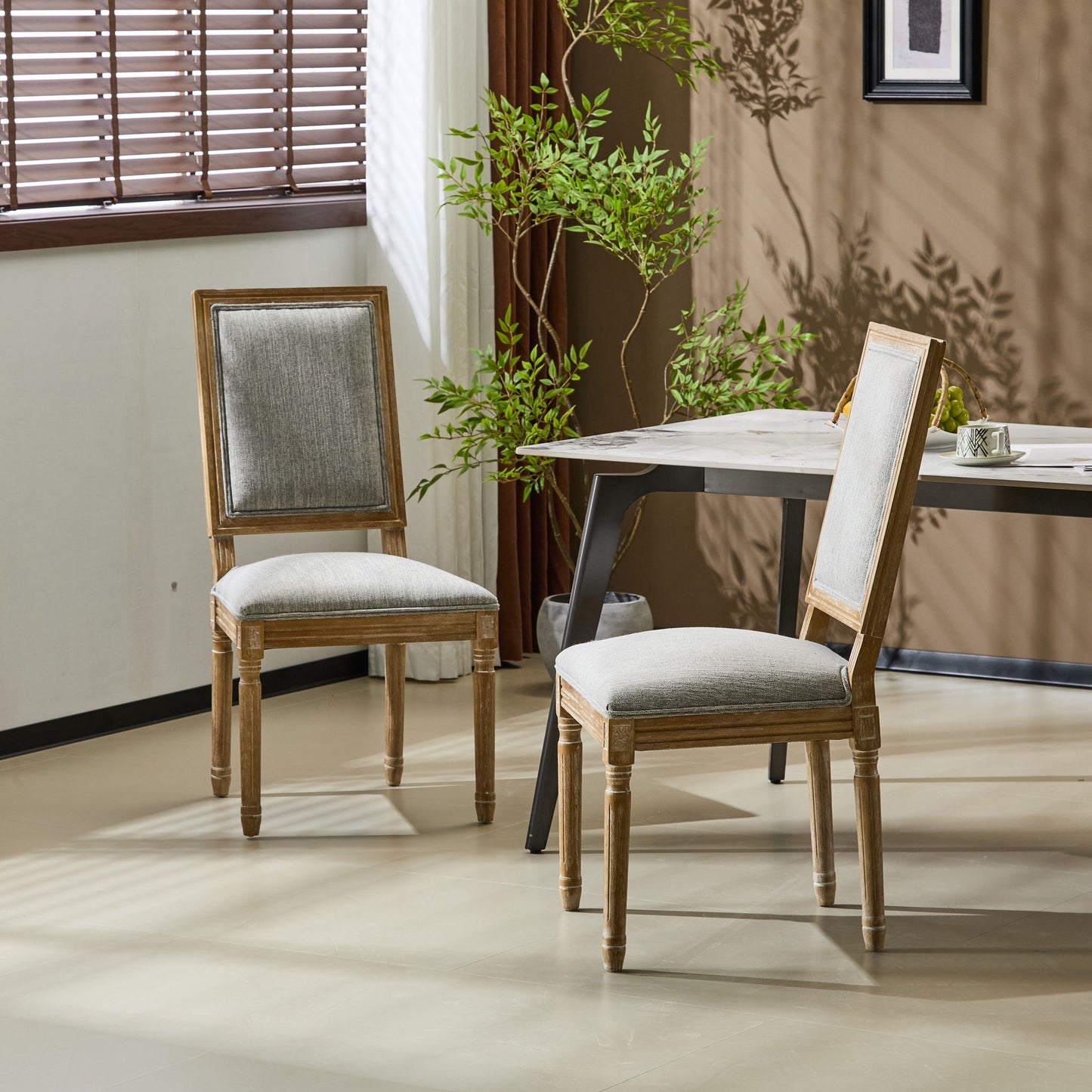 Two French Country Style Dining Chairs with light gray upholstered seats and fluted legs are placed near a modern table in a sunlit room with light walls, a plant, and window blinds casting shadows on the floor and wall.