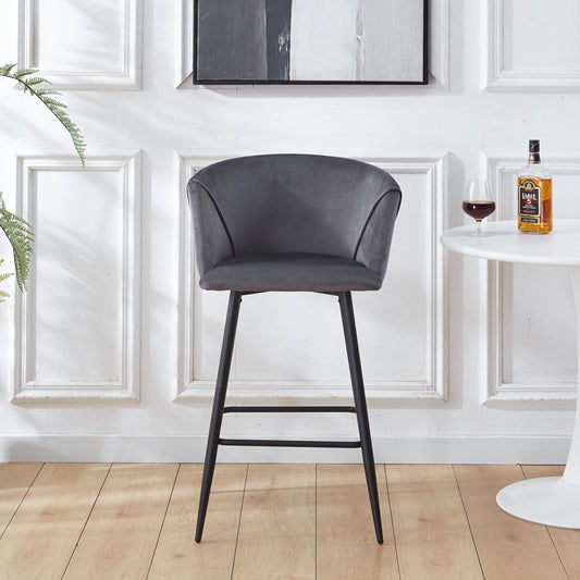 A Counter Height Bar Stool Metal Legs (Set of 2) with curved gray upholstered seats and black metal legs stands on a wooden floor beside a white round table holding a bottle and glass, against a wall with decorative molding.