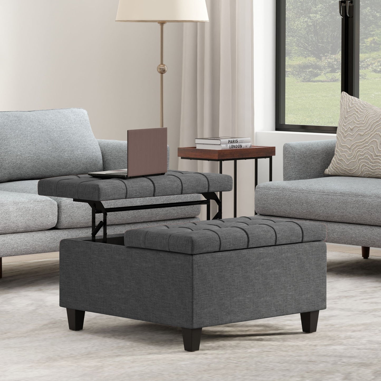 The Harrison Upholstered Coffee Table Storage Ottoman with a lift-top tray sits between two light gray sofas in a modern living room. Nearby, a table with books and a lamp stands beside a large window with white curtains.