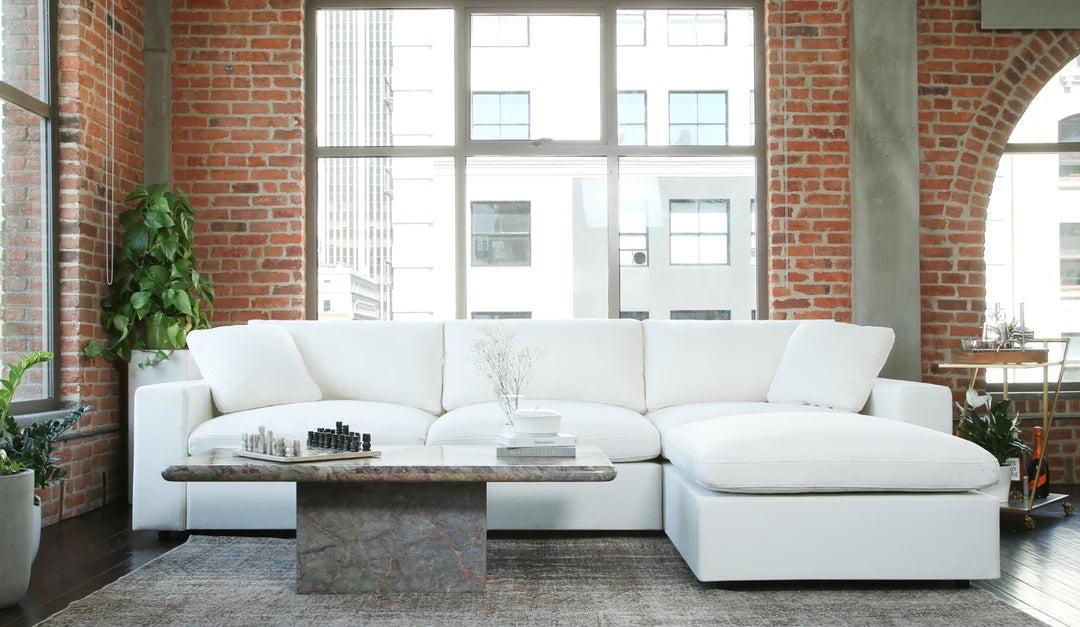 A spacious, modern living room with a large white sectional sofa against a brick wall. A low rectangular coffee table in front of the sofa displays a chess set and vase. Tall windows allow natural light to flood in, highlighting industrial and contemporary elements.