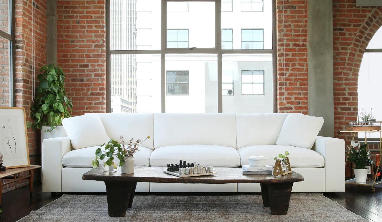 A modern living room with exposed brick walls and large windows. A white sofa is centered against the wall, and a rustic wooden coffee table with plants, a chess set, and decorative items is in front of it. Natural light fills the space, creating a cozy ambiance.