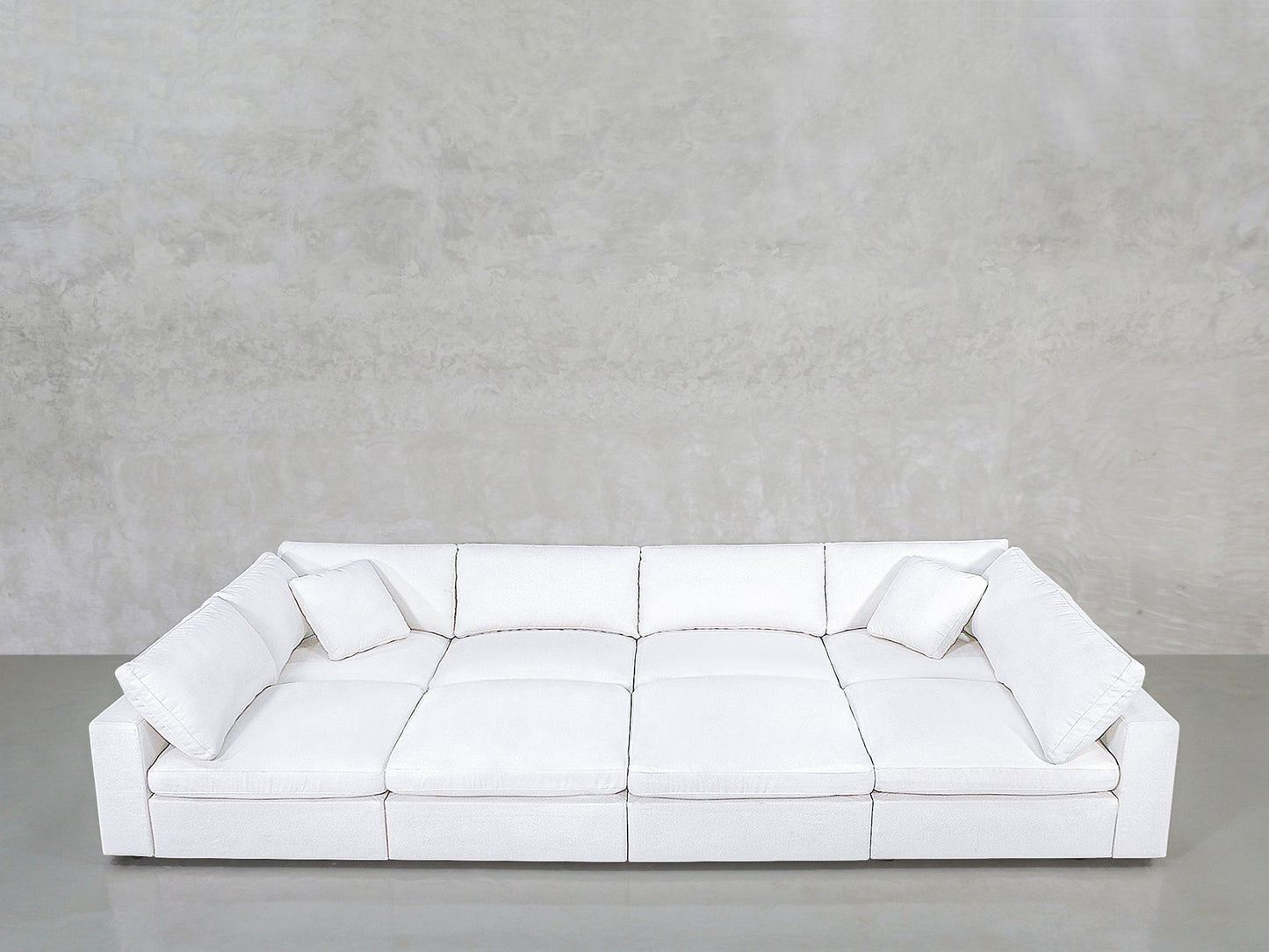 The 8-Seat Modular Pit is a large, modern white sofa featuring wide seats, square armrests, and two matching white cushions, set against a plain light gray wall and smooth concrete floor.