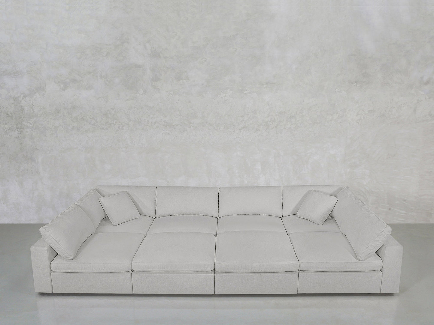 The 8-Seat Modular Pit is a modern, light gray, low-profile sofa with wide armrests and two matching square cushions, shown against a textured light gray wall and smooth gray floor.