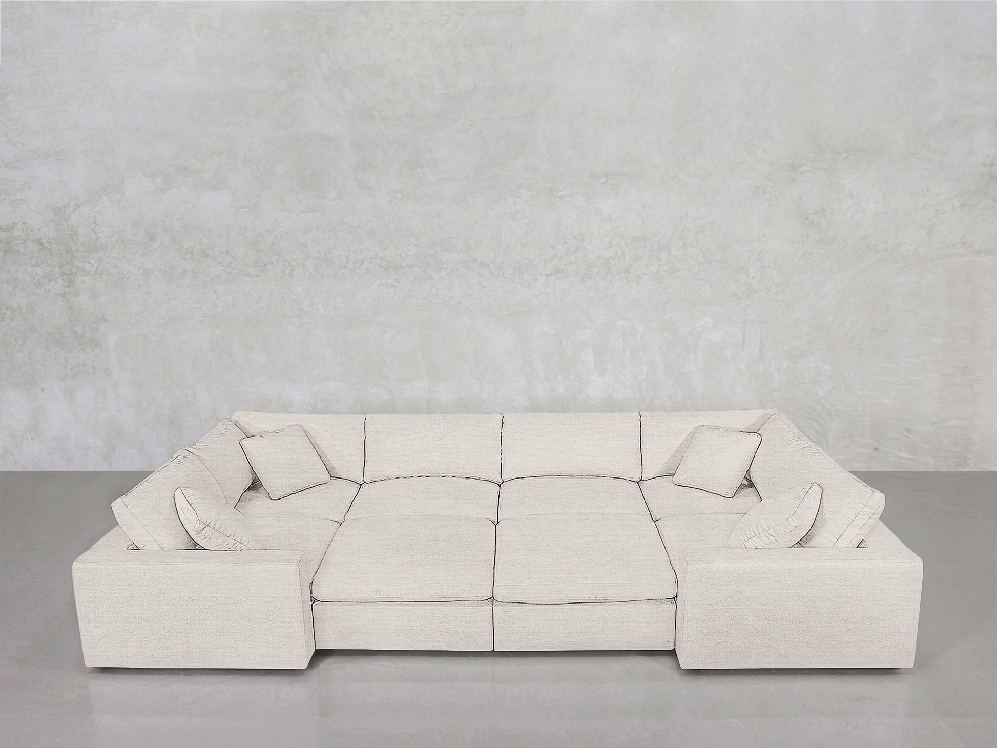 The 8-Seat Modular Closed Pit, a spacious light beige sectional with four matching cushions, is arranged against a plain light grey concrete wall on a smooth grey floor.