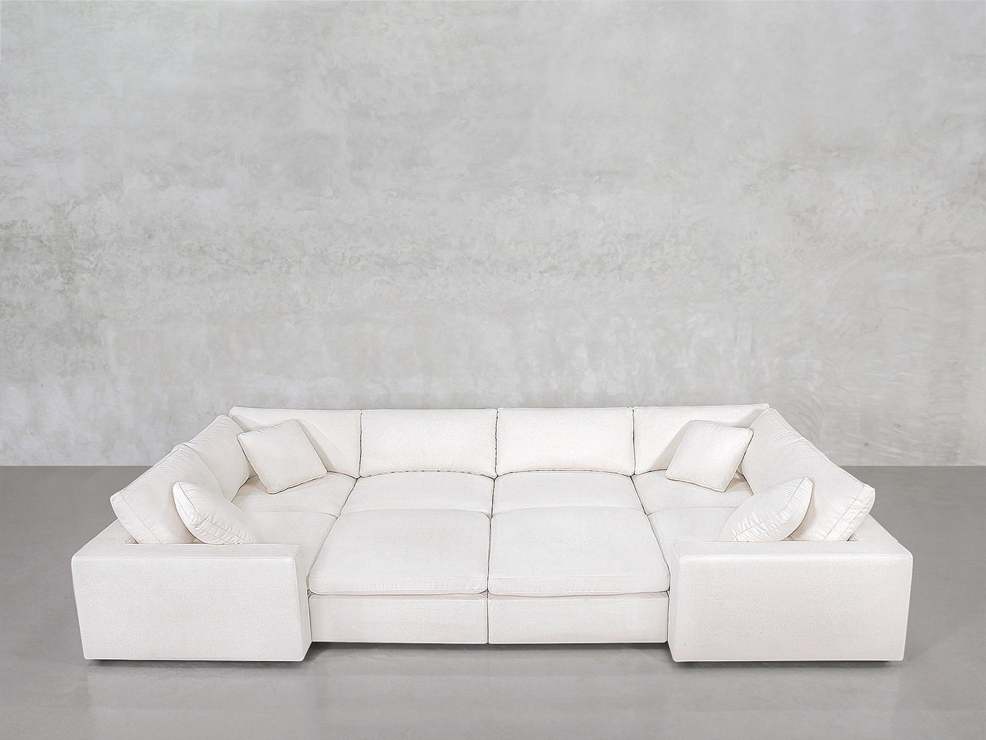 The 8-Seat Modular Closed Pit, a large white U-shaped sectional with six matching throw pillows, is arranged on a gray concrete floor against a light gray textured wall.