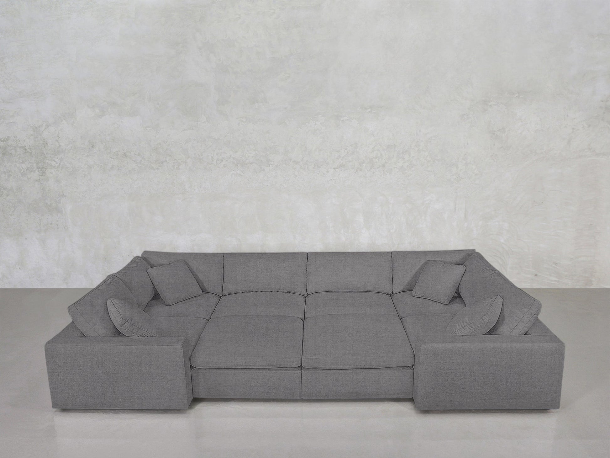 The 8-Seat Modular Closed Pit is a large, modern gray sectional with wide armrests, four matching cushions, and a minimalist design, set against a light gray textured wall and smooth gray floor.