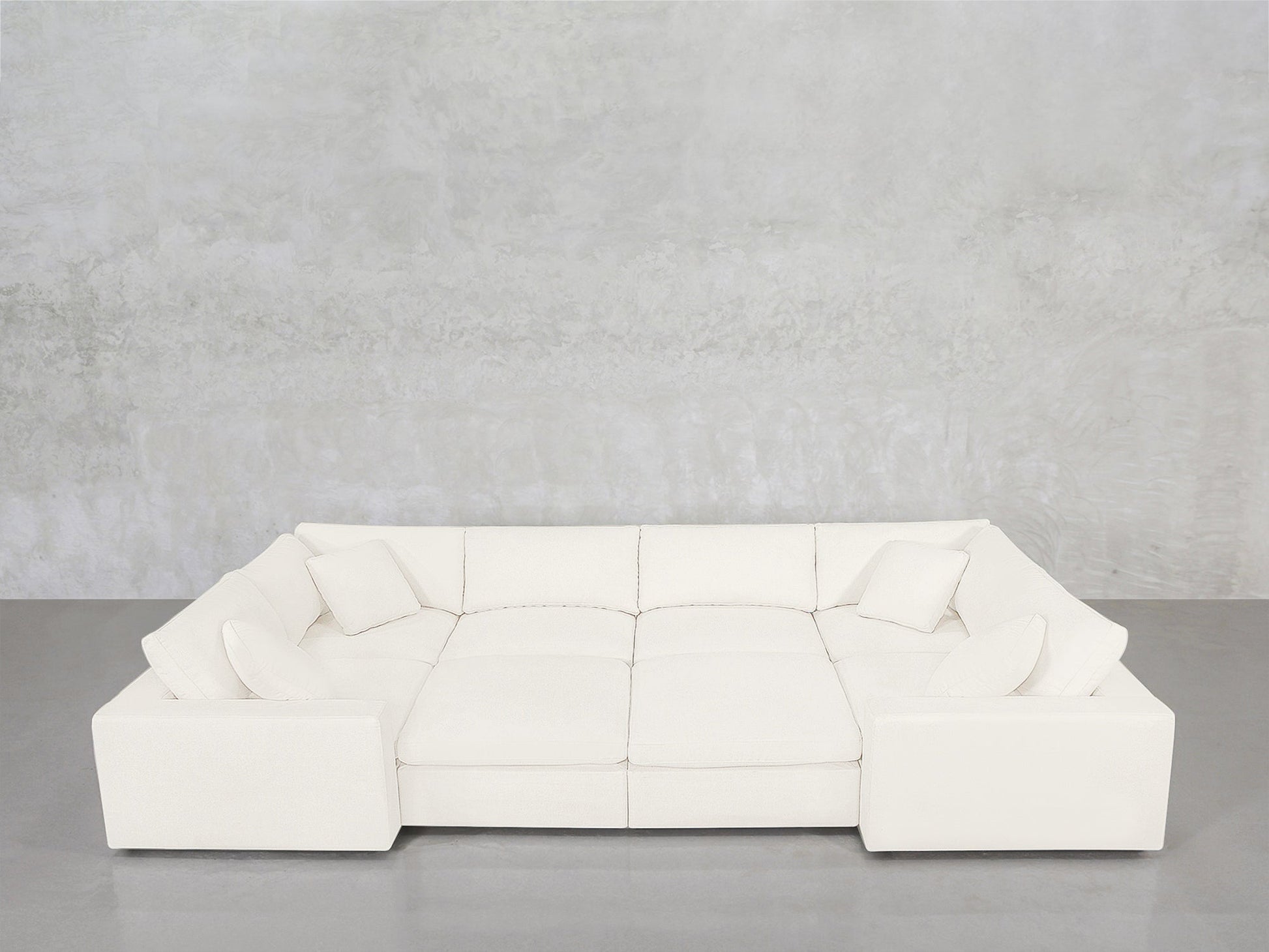 An 8-Seat Modular Closed Pit sectional in modern white, featuring four seat cushions and six matching pillows, is arranged against a light gray concrete wall and a gray floor.