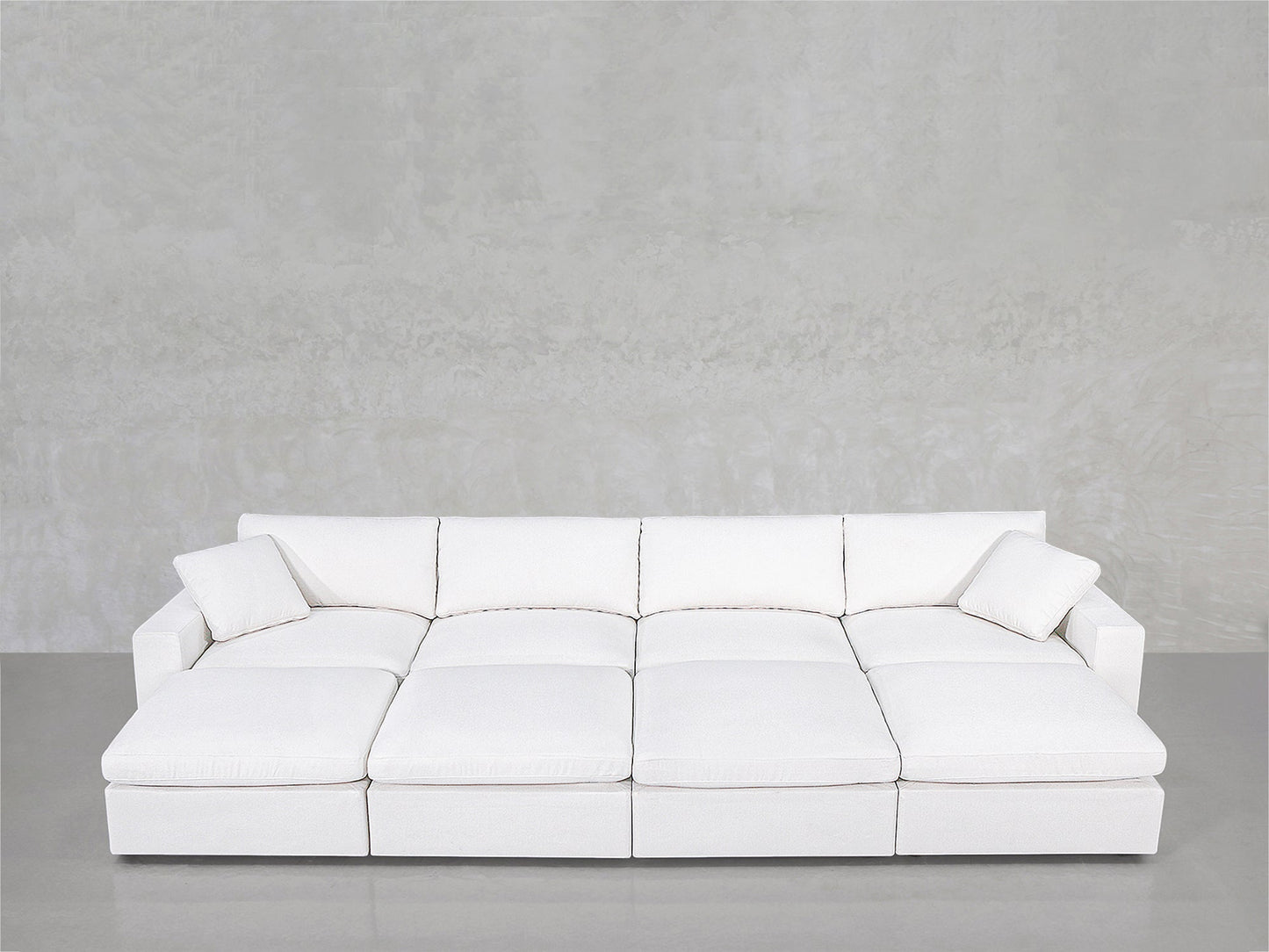 The 8-Seat Modular Daybed features a modern white design with four seat cushions and two matching throw pillows, set against a light gray textured wall and smooth gray floor.