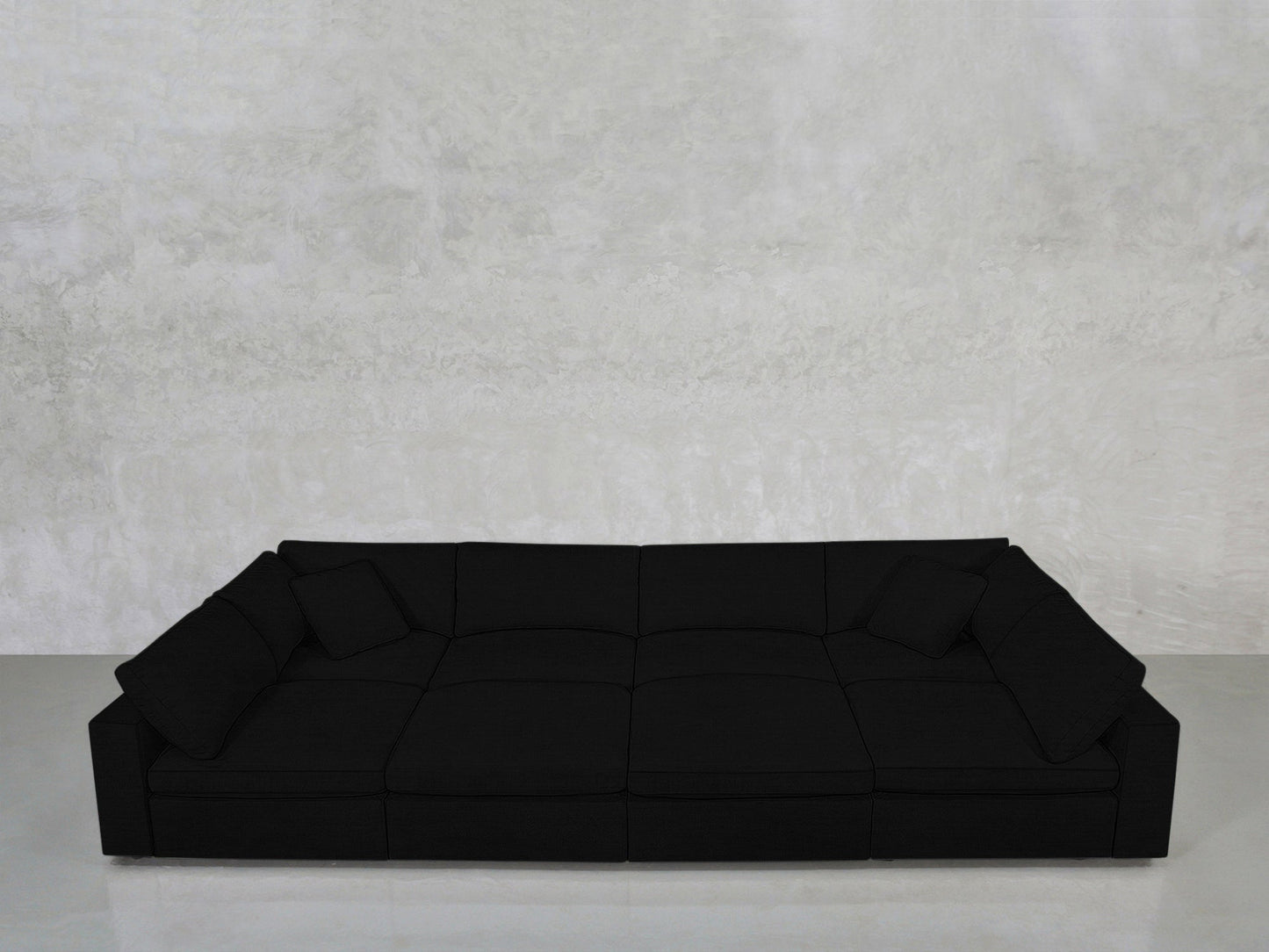 An 8-Seat Modular Pit in modern black with low backrests and three cushions sits against a light gray textured wall on a smooth, light-colored floor.