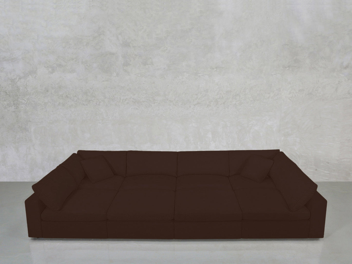 The 8-Seat Modular Pit, a large dark brown sectional with cushions, is set against a textured light gray concrete wall on a light-colored floor.