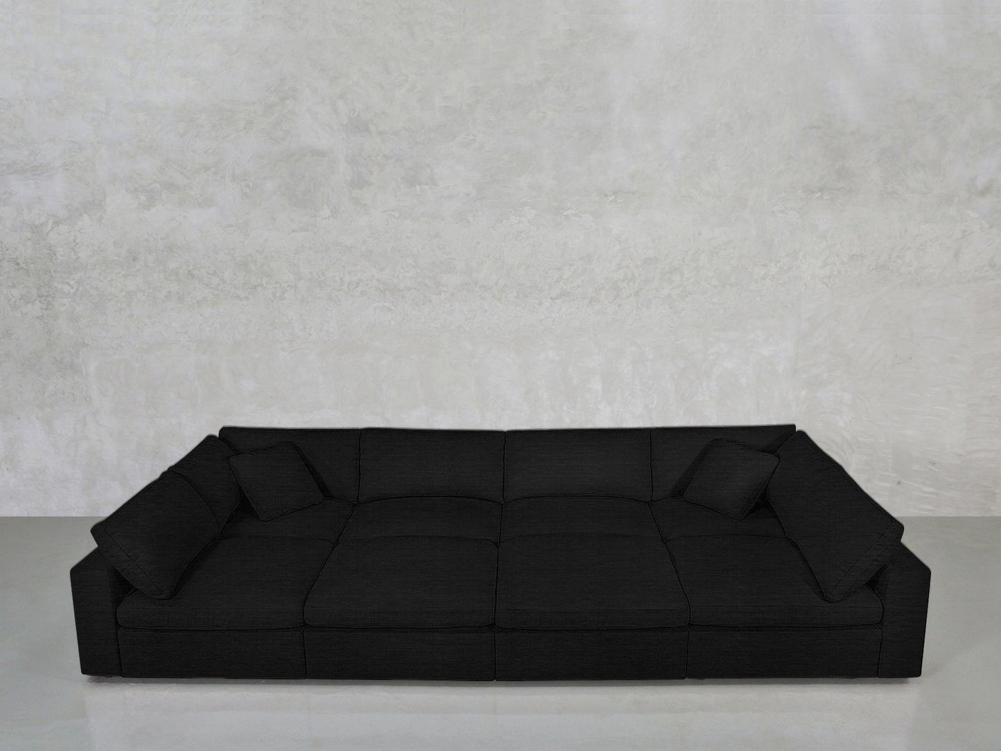 The 8-Seat Modular Pit is a modern, low-profile black sofa with wide seats and two matching black cushions, set against a light gray textured wall on a smooth, light-colored floor.