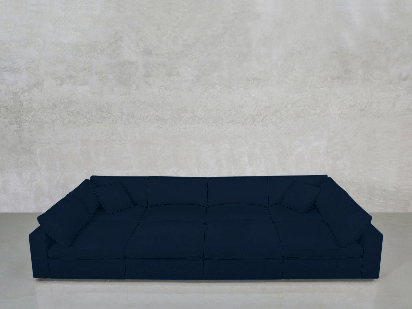 An 8-Seat Modular Pit sectional in dark blue with wide seats and low backrests is set against a textured light gray concrete wall and smooth gray floor, with two matching blue cushions placed at each end.