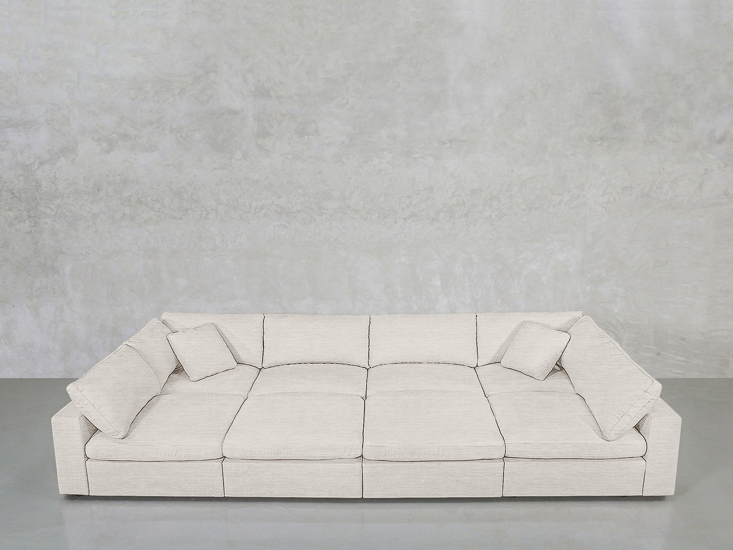 An 8-Seat Modular Pit in light gray, featuring three seat cushions and four matching throw pillows, sits against a textured light gray wall on a smooth gray floor.