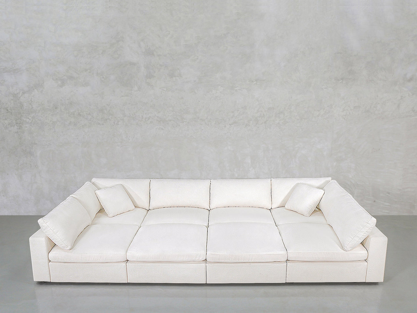 The 8-Seat Modular Pit is a large, modern white sectional with wide cushions and two matching throw pillows, set against a light gray textured wall and smooth gray floor.