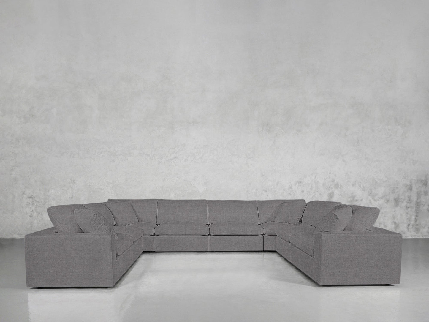 An 8-Seat Modular U-Sectional with a modern gray design and matching cushions is set in a minimalist room with a light gray textured wall and a sleek, shiny floor.