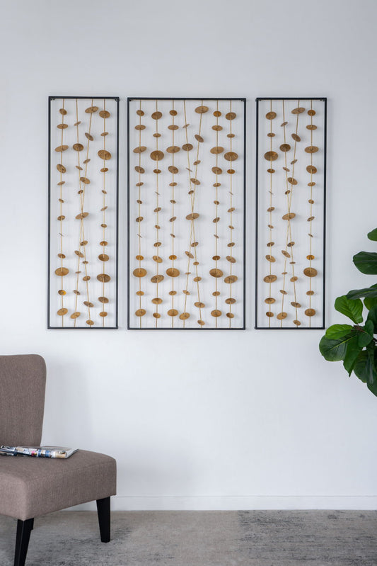 Three Metal Decorative Wall Art With Frame, Wall Decor For Living Room (Set of 3) - Gold panels are mounted on a white wall above a gray upholstered chair with a magazine on it. A green potted plant sits to the right.