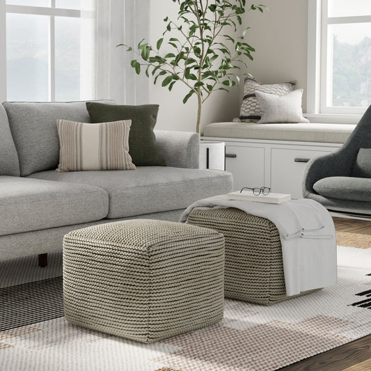A modern living room with a gray sofa, patterned cushions, two Eastmont Outdoor/Indoor Poufs in cream, a cozy window seat, a plant, and a gray armchair. A white blanket and glasses rest on one pouf as sunlight streams through large windows.