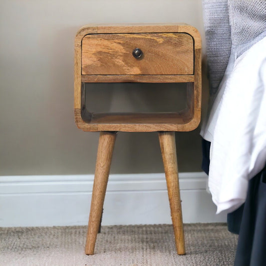 The Mini Chestnut Curve Lower Bedside Nightstand, featuring a small wooden design with rounded edges, a single drawer with knob, and an open shelf below, stands next to a bed on a carpeted floor.