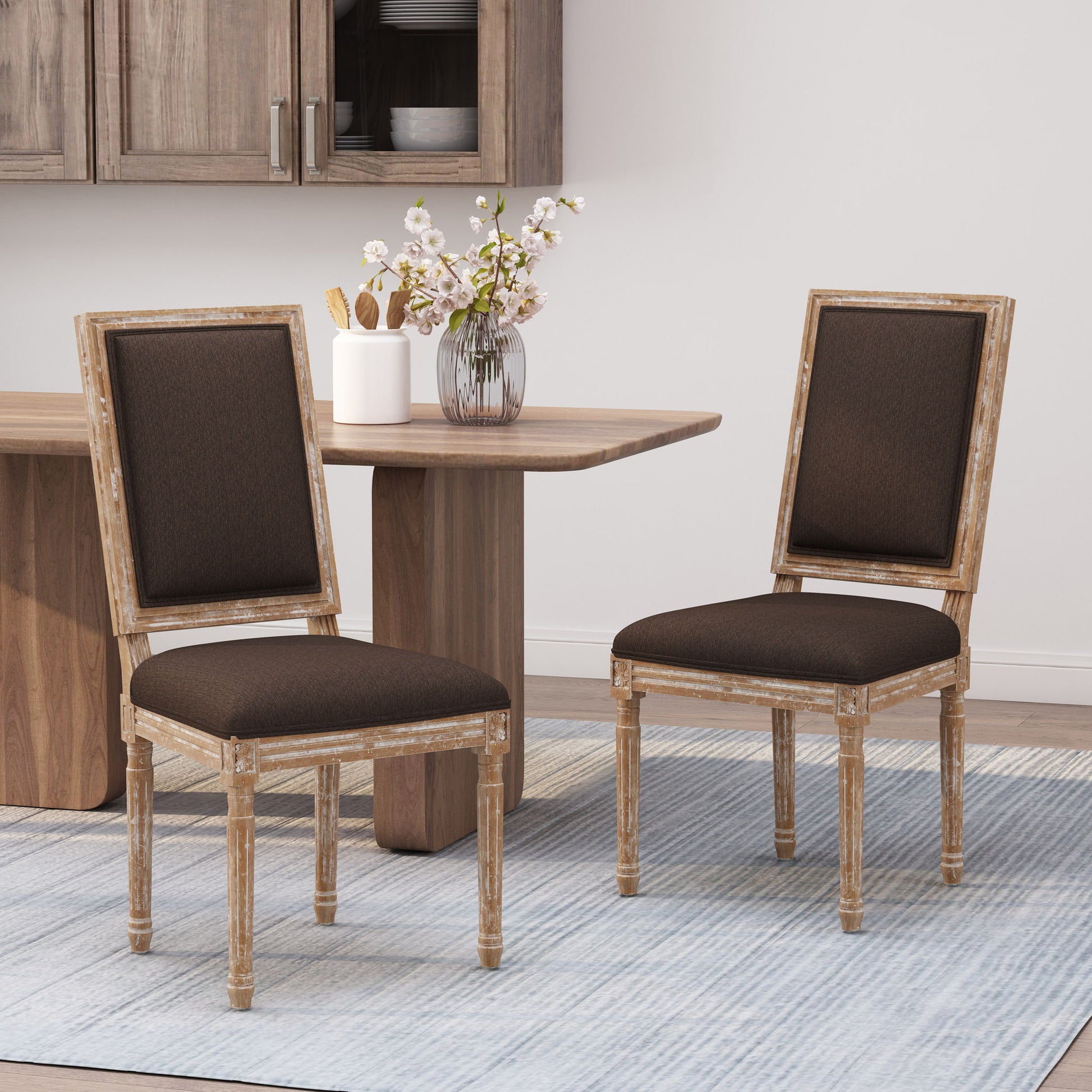Two French Country Style Dining Chairs with upholstered seats and fluted legs sit next to a wooden table in a modern dining space, with floral vase decor and kitchen cabinets visible in the background.