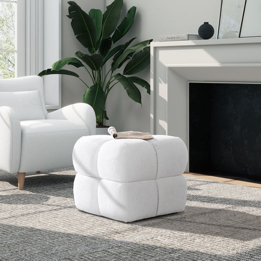 A modern living room features the Pixel Oversized Square Tufted Pouf Ottoman in Almond White at the center, a white armchair to the left, fireplace to the right, lush green plant behind, and sunlight streaming through a nearby window.