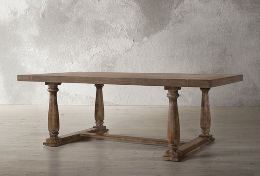 The Bernard - Weathered Dining Table features a thick rectangular wooden top and four carved pedestal legs joined by a horizontal base, shown against a textured gray wall and light wood floor.
