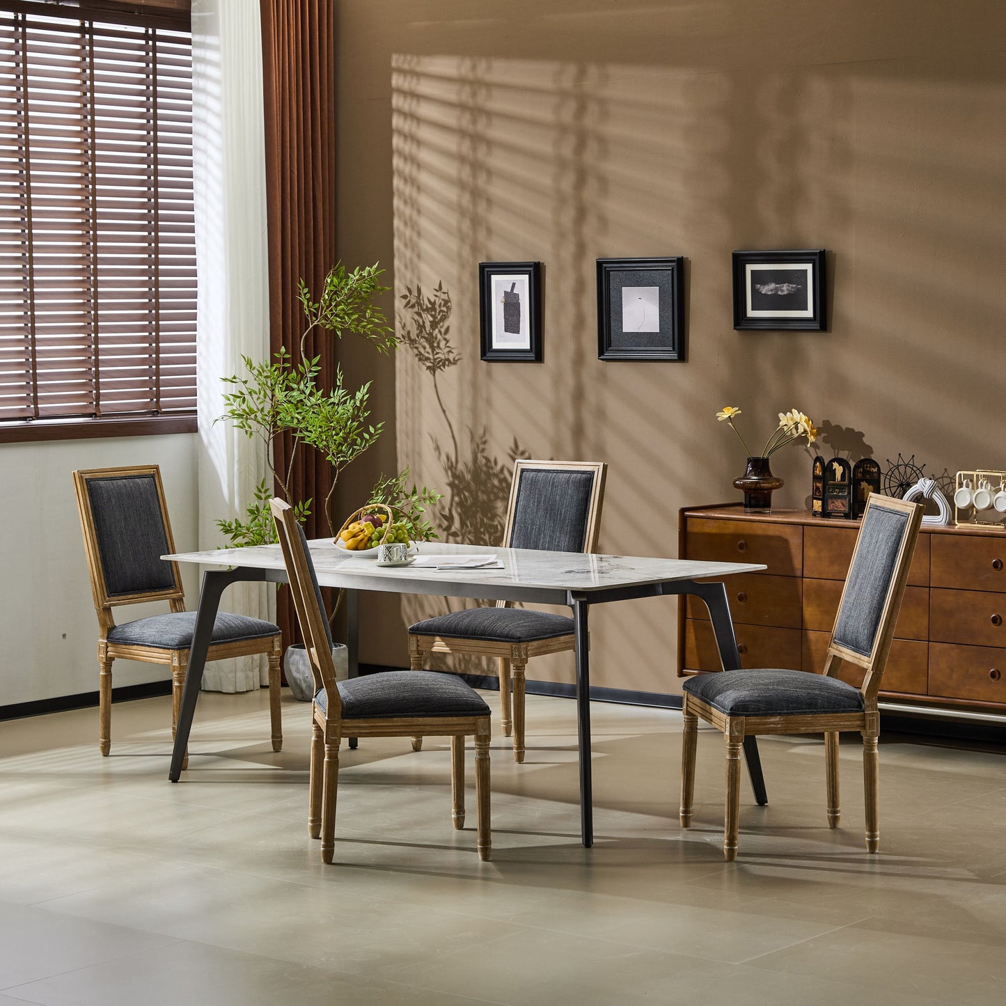A modern dining room with a rectangular table, four French Country Style Dining Chairs with upholstered seating and fluted legs, a wooden sideboard, wall art, a potted plant, and sunlight streaming through window blinds.