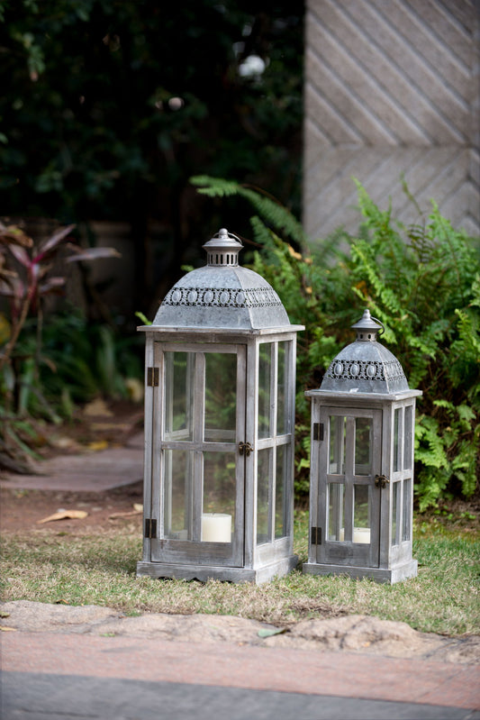 Two gray Wooden Candle Lantern Decorative hurricane holders of different sizes are placed on outdoor grass, each with a white candle inside. Ferns and plants are visible in the background. Perfect for home, garden, or wedding decor indoors or outdoors.