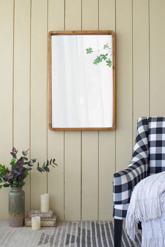 The Rectangle Wood Mirror, Farmhouse Wall Decor in brown hangs on a beige paneled wall. Nearby are a checkered armchair with a white throw, two books, a vase of greenery, candles, and the mirror reflects leafy branches.