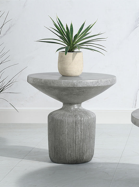 The Laddie - Weathered Table is a modern round concrete side table with a textured pedestal base, shown with a small potted green plant on top against a light-colored wall and floor.